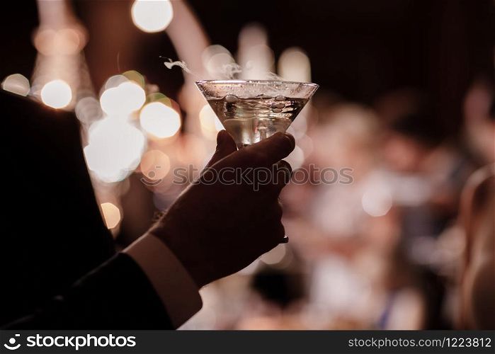 A close up of a man&rsquo;s hand who is dressed in formal attire and holding a martini glass on party background. A close up of a man&rsquo;s hand who is dressed in formal attire and holding a martini glass on party background.