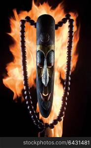 A close-up of an ancient wooden mask with beads against fire flames.