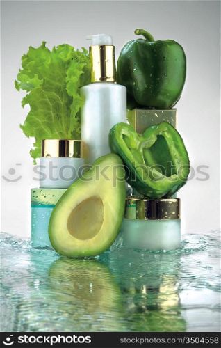 A close-up of different jars of cosmetics and green vegetables in water splash.