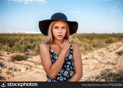 a close-up portrait of a blue-eyed blonde girl in a black hat and a floral print dress.she&rsquo;s posing in a vineyard during sunset