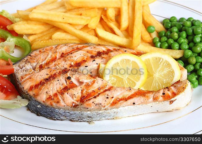 A close-up view of a grilled salmon steak served with salad, chips, peas and lemon slices.