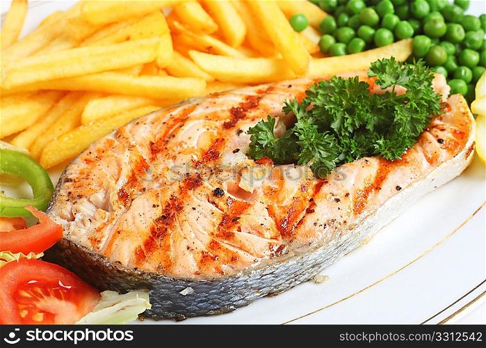 A close-up view of a grilled salmon steak served with salad, chips, peas and lemon slices.