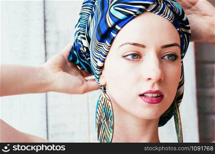 a closeup portrait of a pretty young woman wearing a colorful blue headscarf in an african manner, and big round earrings, blue eyes, light background