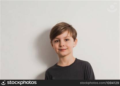a closeup portrait of a smiling cute boy in a grey shirt, leaning on the white wall