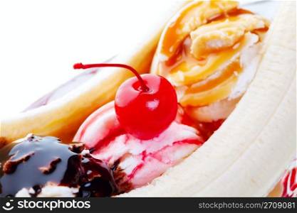 A closeup view of a delectable banana split, with chocolate, strawberry, and caramel drizzled on mounds of vanilla ice cream and topped with a red cherry. Shot on white background. Selective focus on cherry.