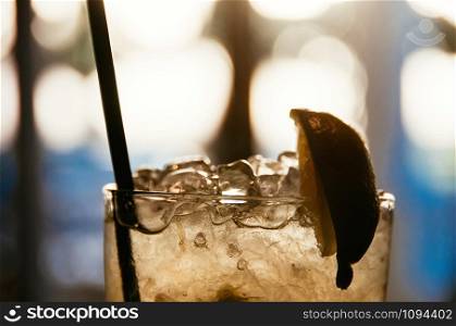 a closeup view of an old fashion glass with cocktail, ice cubes, black sipping straw and lime, blurred bokeh in the background