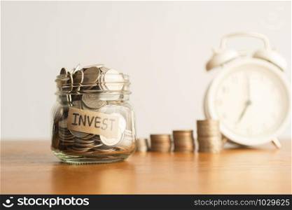 A coin in a glass bottle, a pile of coins and a white retro alarm clock on a brown wooden table. Investment business, retirement, finance and saving money for future concept.