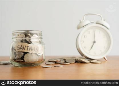 A coin in a glass bottle, a pile of coins and a white retro alarm clock on a brown wooden table. Investment business, retirement, finance and saving money for future concept.