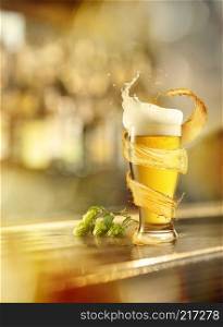 A cold glass of beer with a splash around and with a branch of hops on a wooden background on a blurred background. A cold glass of beer with a splash and with a hops on a wooden background