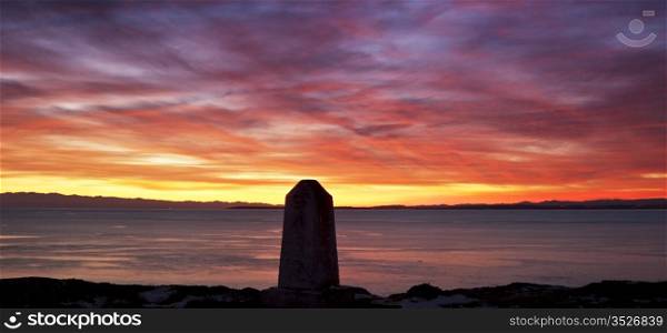 A colorful, dramatic sunset from San Juan island looking west towards the Straits of Juan de Fuca and Vancouver Island. The concrete obelisk in the foreground is a US geological marker.