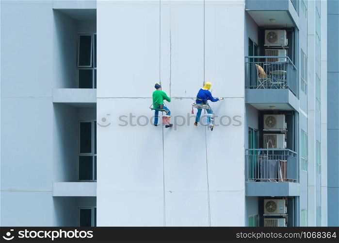 A company group of people, workers or painters, painting a wall on a high rise office building with cable wires outside in Bangkok Downtown, urban city at noon.