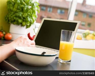 A computer in the kitchen with a bowl of soupr or cereal and orange juice - shallow depth of field with focus on juice and hand