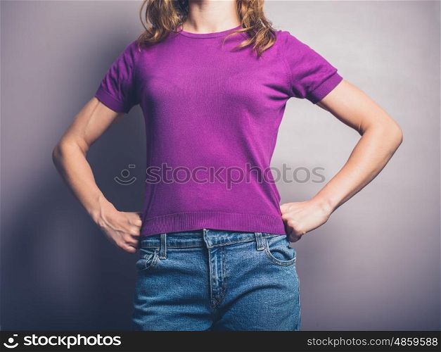 A confident young woman in purple shirt is standing in a powerful pose with her hands on her hips