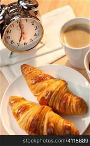 A continental breakfast of croissant pastries and coffee Illuminated with golden early morning sunshine and accompanied by a classic alarm clock set at 7am