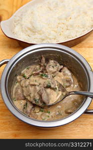 A cooking pot full of beef stroganoff with a serving spoon and a bowl of white rice.