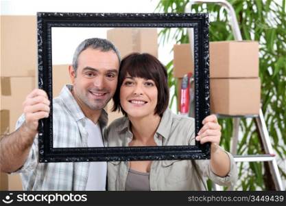 a couple is holding and posing behind a painting frame inside an apartment full of unpacked packages