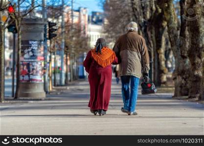 A couple of eldery people in hands are walking through the city street. Rear view.