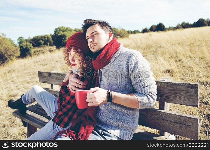 A couple of happy young millennials in love sunbathing on a wooden bench and taking a cup of coffee or tea while they are hugging with the backlight from autumn sun