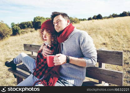 A couple of happy young millennials in love sunbathing on a wooden bench and taking a cup of coffee or tea while they are hugging with the backlight from autumn sun