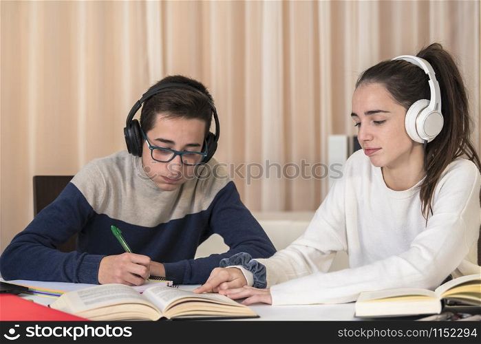 A couple of tired looking teenagers read a book and take notes while listening to a lesson on their headphones on an out of focus background. Student concept.. Teenagers look tired while taking a lesson
