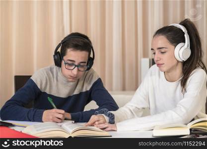 A couple of tired looking teenagers read a book and take notes while listening to a lesson on their headphones on an out of focus background. Student concept.. Teenagers look tired while taking a lesson