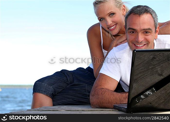 a couple on a pontoon near the sea, the man is doing computer,