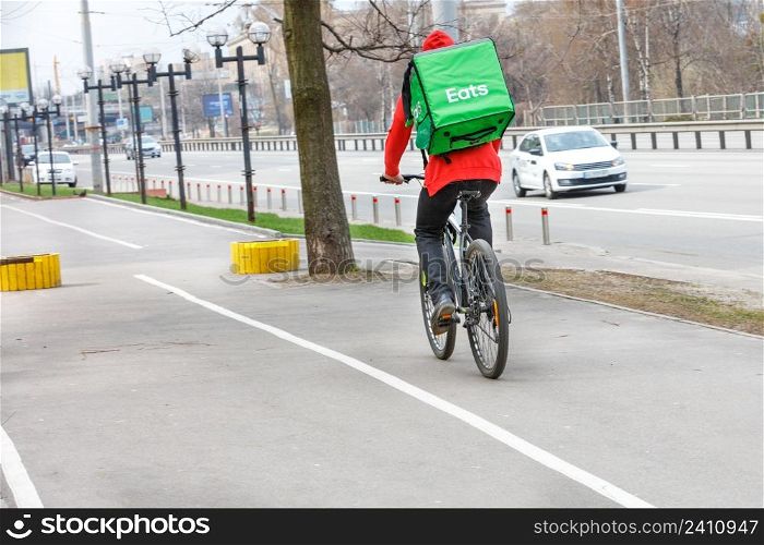 A courier with a green isothermal backpack behind his back rides along the sidewalk of a city street on a bicycle. Modern food delivery concept. Cpoy space.. Courier with an isothermal green backpack on his back on a food delivery bike on a city street.