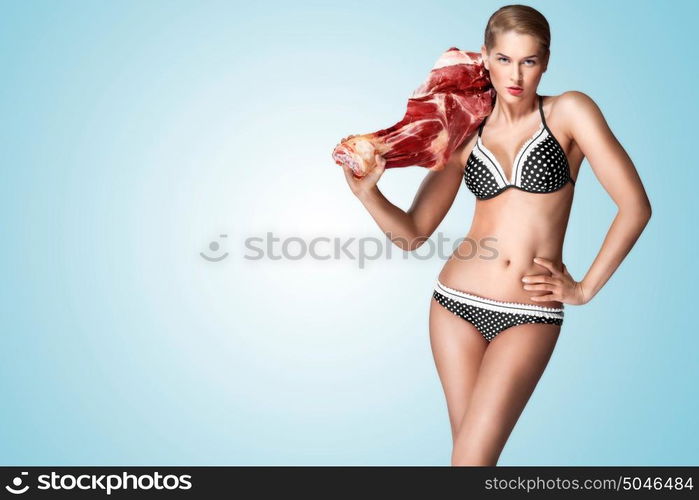 A creative portrait of a beautiful girl in bikini holding raw meat on her shoulder.