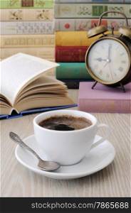 A cup of coffee on the table against the background of an open book with a clock and a stack of books