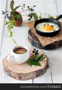 A cup of espresso on a wooden stand in the foreground. Fried chicken egg in the pan for breakfast. Healthy organic foods. Close-up. White vintage wood background. Decorated with leaves and fruits of wild grapes.