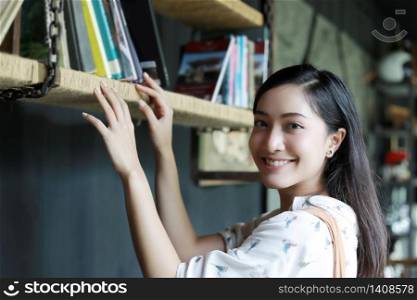 A cute Asian woman choose a book on the bookshelf for reading and she smiled happy on her holiday at home.