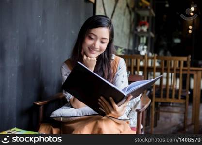 A cute Asian woman is sitting reading a book on the sofa or a chair happy smiling at home during relax time