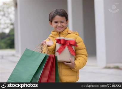 A cute boy in winter coat holding gift box with shopping bag and smiling. preparing for Christmas holiday