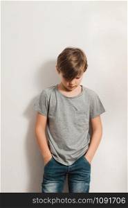 a cute boy stands next to white wall, grey t-shirt, blue jeans, hands in pockets, bowing his head down
