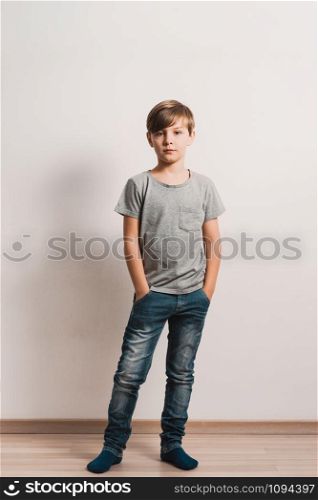 a cute boy stands next to white wall, grey t-shirt, blue jeans, hands in pockets
