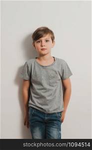 a cute boy stands next to white wall, grey t-shirt, blue jeans, frightened face