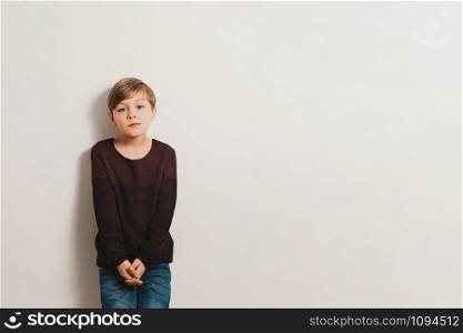 a cute boy stands next to white wall, vinous pullover, blue jeans, unhappy face, copy space