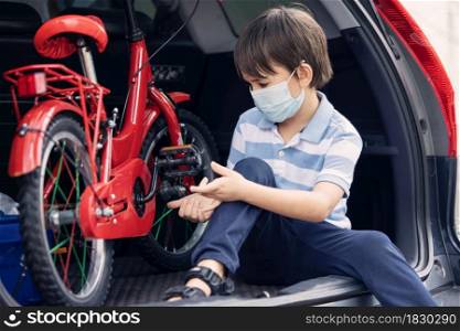 A cute boy wearing face mask sitting in rear car with red bicyle preparing to ride at outdoor during pandemic of coronavirus covid 19.Social distance.