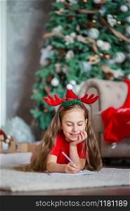 A cute girl in a Santa hat writes a letter to Santa near the Christmas tree. Happy childhood, a time for fulfilling desires. Merry Christmas.. A cute girl in a Santa hat writes a letter to Santa near the Christmas tree. Happy childhood, a time for fulfilling desires.