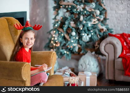 A cute girl in a Santa hat writes a letter to Santa near the Christmas tree. Happy childhood, a time for fulfilling desires. Merry Christmas.. A cute girl in a Santa hat writes a letter to Santa near the Christmas tree. Happy childhood, a time for fulfilling desires.