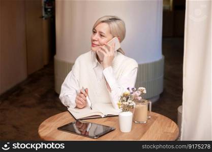 A cute girl is sitting in a cafe and talking on the phone and making notes in a diary, there is a phone and a tablet nearby. A cute girl is sitting in a cafe and talking on the phone and making notes in a diary