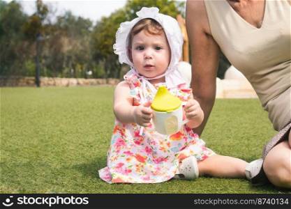 A cute little baby girl toddler wearing a white bonnet and a dress sat on green grass with her grandmother and holding a sippy cup with water