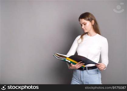 A cute student girl looks into an open folder for documents and stands on a gray background.. A cute student girl looks into an open folder for documents and stands on a gray background