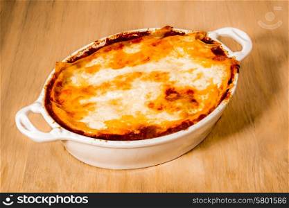 A delicious looking dish of lasagna with a golden yellow melted layer of cheese on top, inside a white ceramic dish, placed on a wooden surface.
