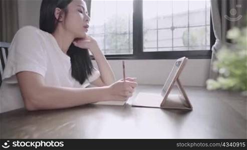 A demotivated attractive asian student sitting on a living room, hand on chin, young bored girl writing on a paper book, home e-learning, people and distant educational technology, self isolation