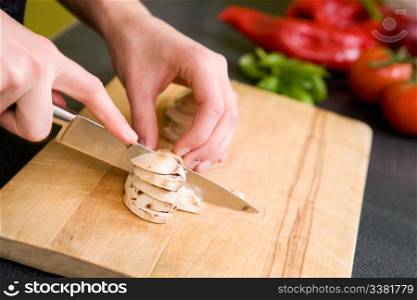 A detail image of a female hand slicing mushrooms for a pizza. Focus is on the hand and mushrooms while tomatoes and peppers are out of focus in the background.