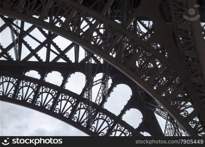 A detail view of a portion of one of the base arches on the Eiffel tower that shows details of the art deco ironwork silhouetted against a cloudy sky..