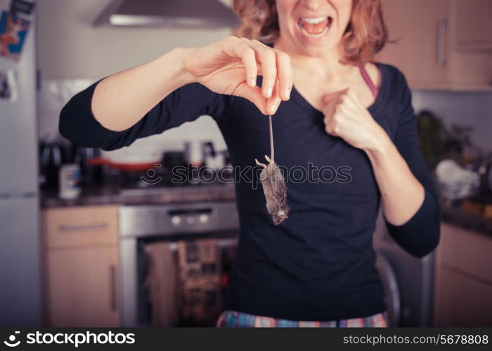 A disgusted young woman is holding a dead mouse by it&rsquo;s tail in the kitchen