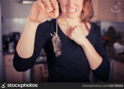 A disgusted young woman is holding a dead mouse by it&rsquo;s tail in the kitchen