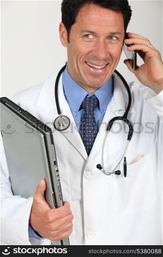 A doctor holding a laptop and having a conversation via his mobile phone.